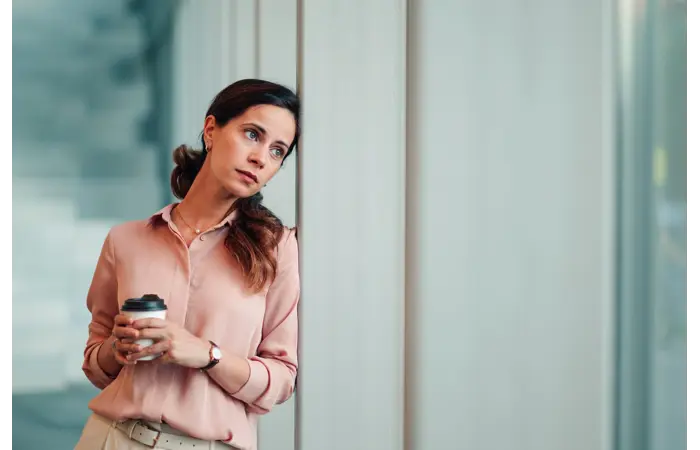 Woman standing alone beside a glass wall, looking thoughtful while holding a coffee