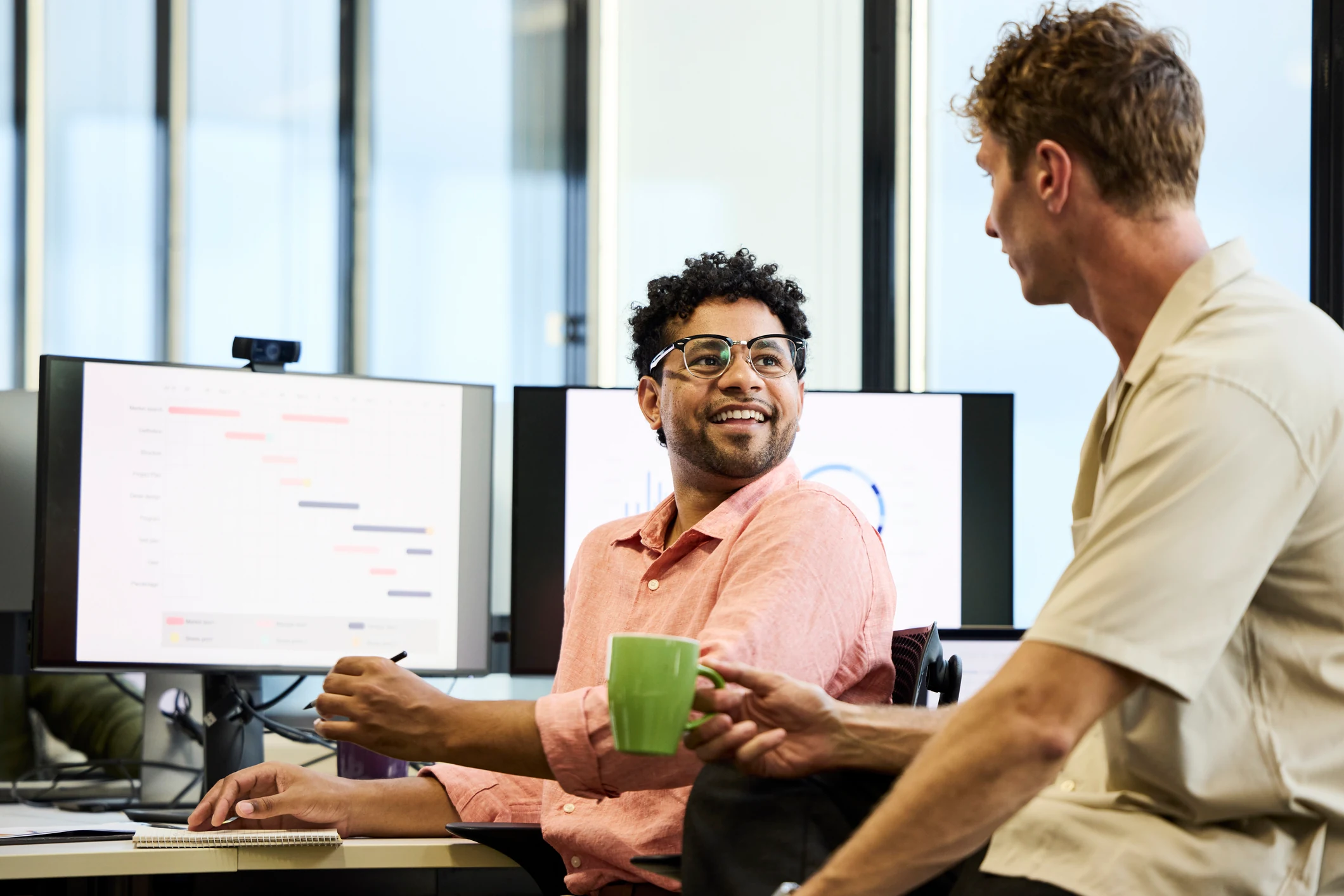 Man listening to his colleague while sat at his desk and smiling warmly.
